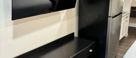 Una cocina moderna con un televisor de pantalla plana montado en la pared, una encimera negra con cajones, un refrigerador de acero inoxidable y armarios blancos.