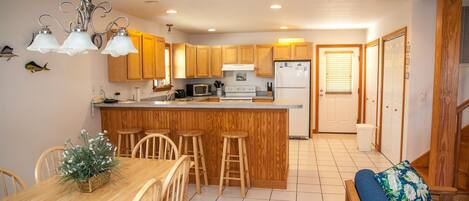 A kitchen with wooden cabinets, a central island with stools, a dining table with chairs, and a blue sofa with a floral pillow.