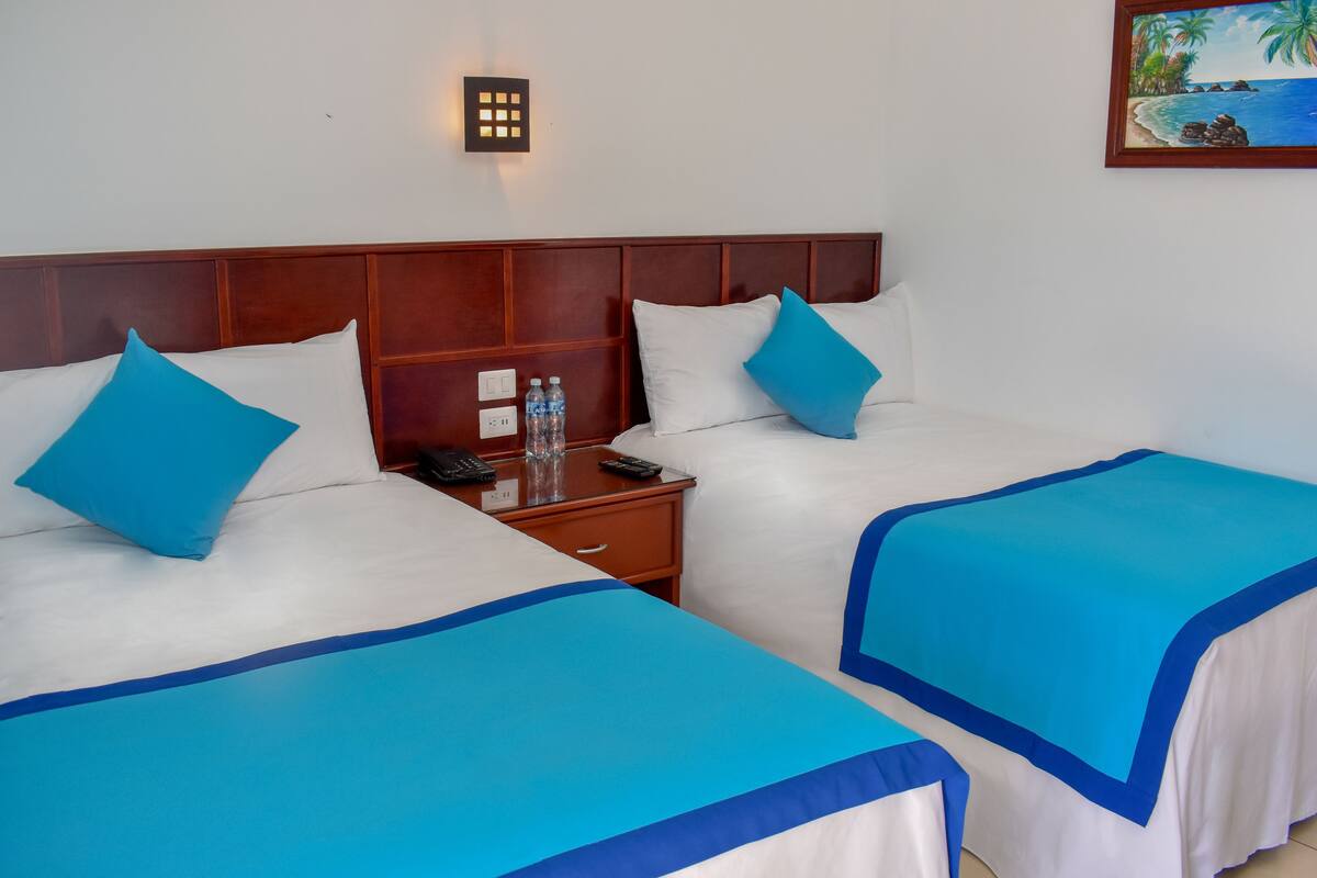 Quarto de hotel com duas camas, cada uma com roupa de cama azul e branca, cabeceira de madeira, mesa de cabeceira com telefone e controles remotos, e luminária fixada na parede.