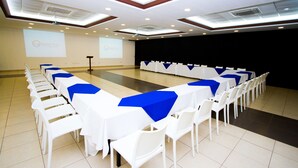 A conference room with rows of white tables and chairs, a stage with a projector screen, and a dark screen wall.
