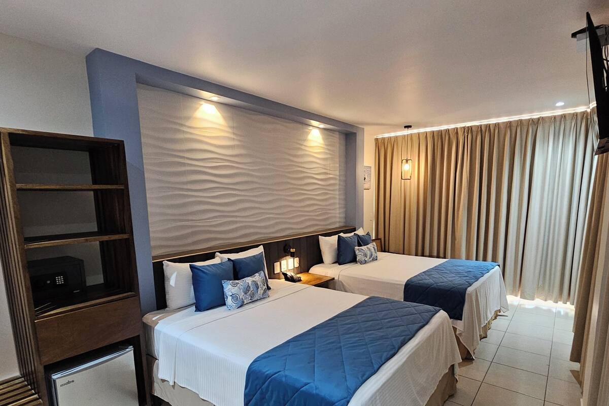 Quarto de hotel com duas camas, um minibar, uma escrivaninha e uma janela grande com cortinas.