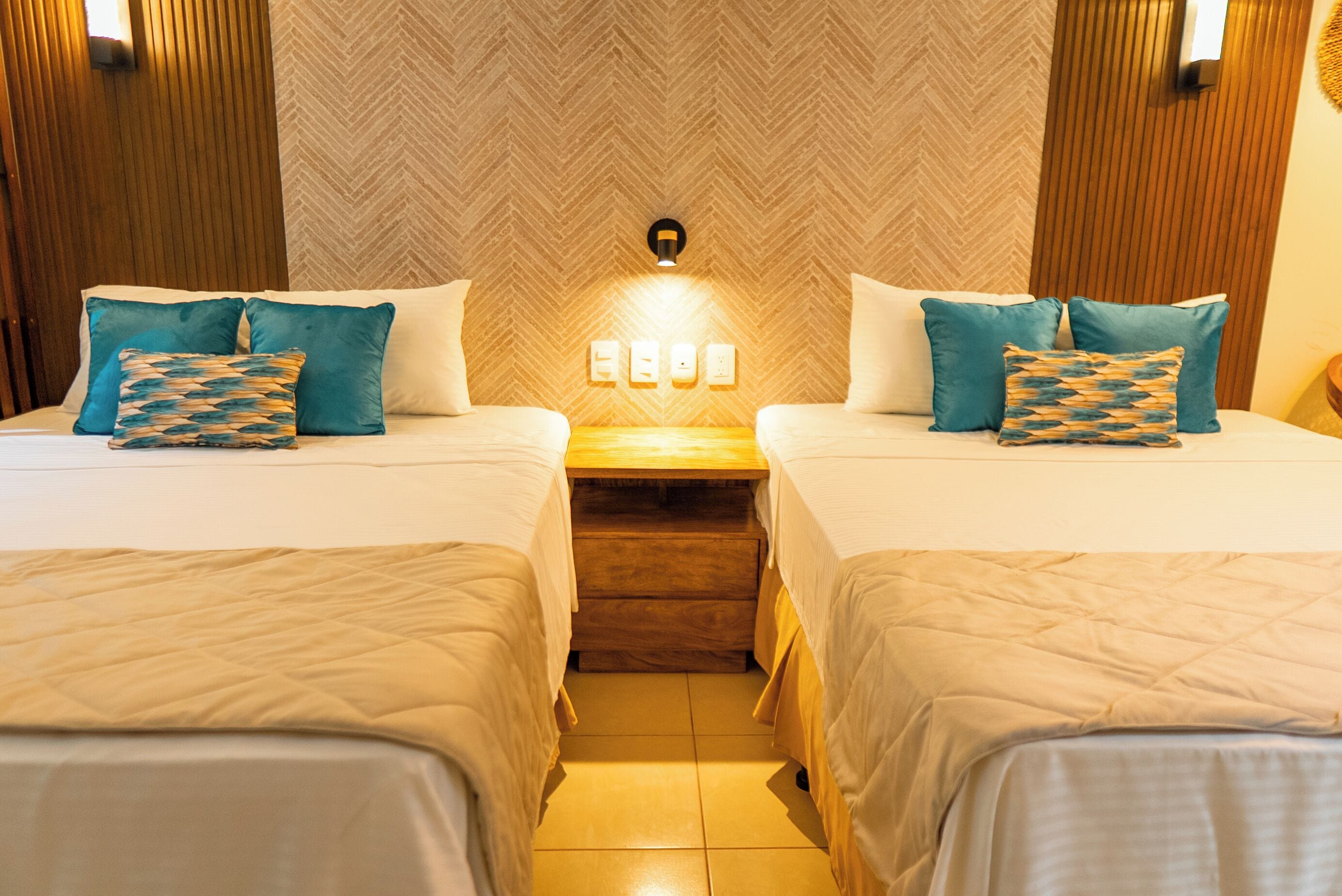 A hotel room with two beds, wooden headboard panels, a herringbone-patterned wall, and a nightstand with a wall-mounted light.