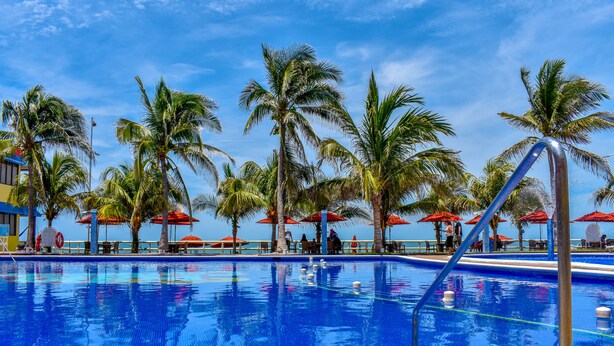 A swimming pool with palm trees and red umbrellas, surrounded by a poolside area with chairs and tables.