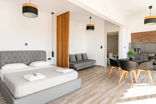 A modern hotel room with a large bed, a gray sofa, a dining area with a table and chairs, and a kitchen in the background.