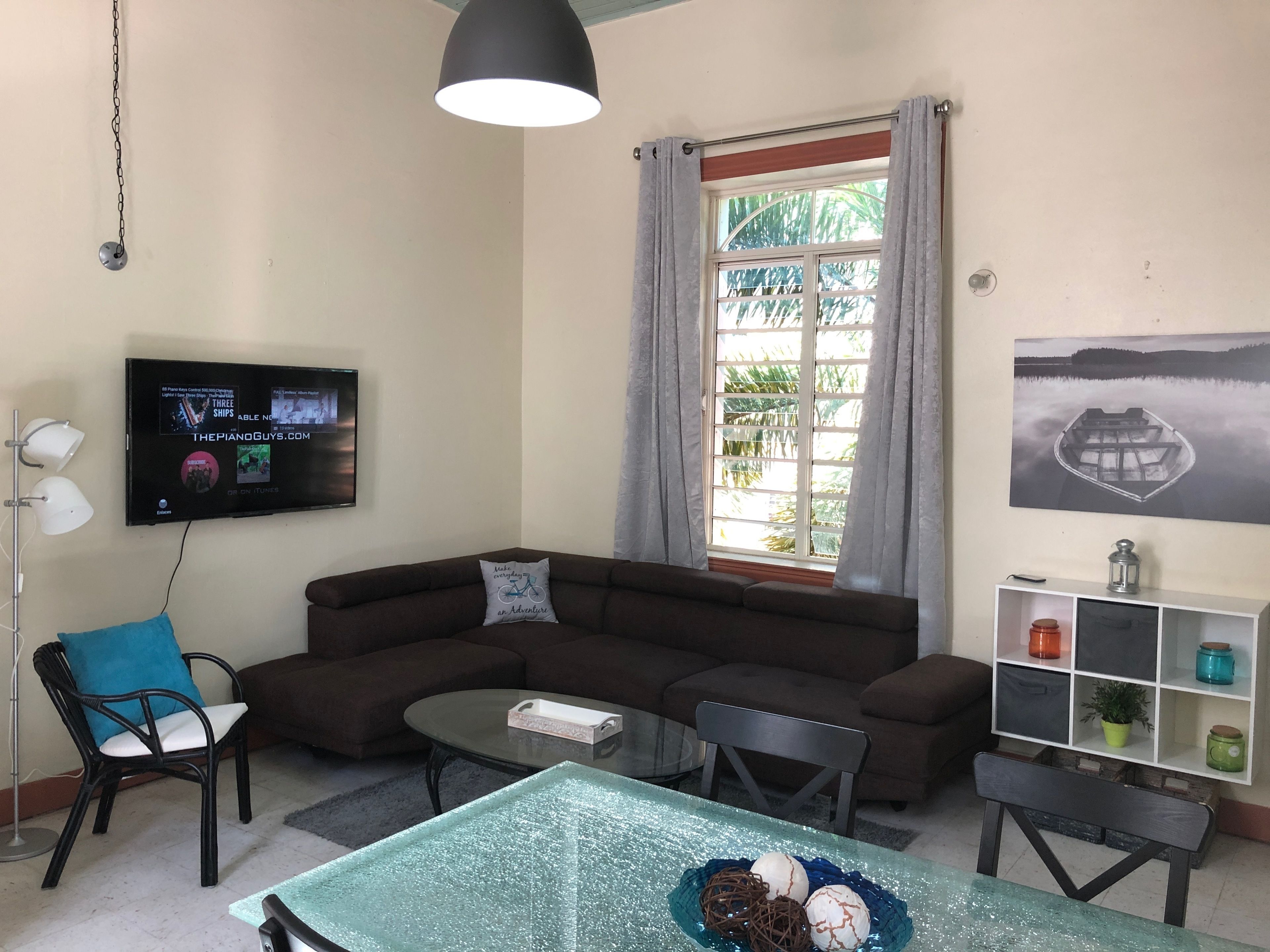 A living room with a sectional sofa, a glass-top coffee table, a TV mounted on the wall, and a window with curtains.