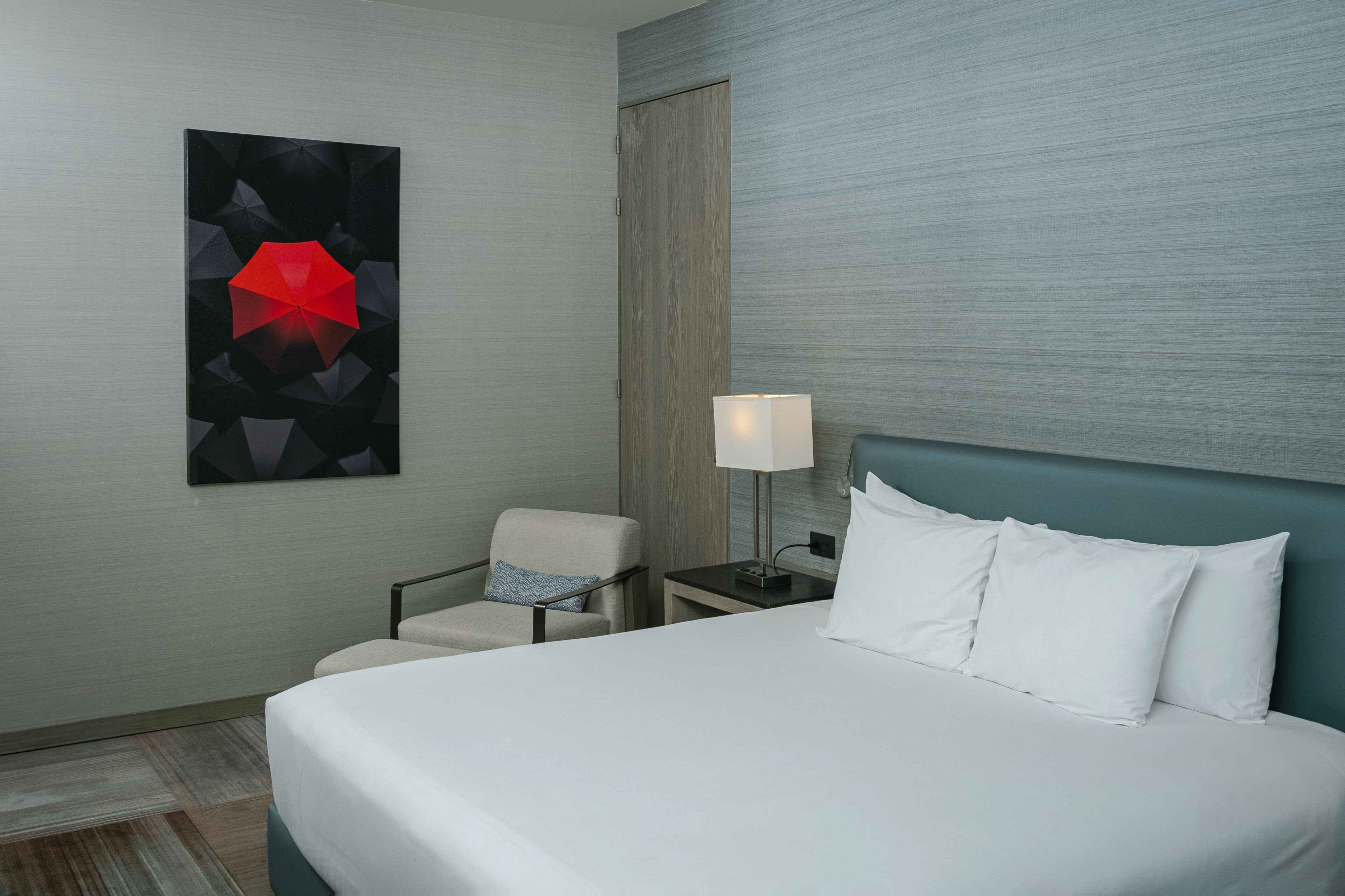 Habitación de hotel con cama, una silla, una mesita de noche con lámpara y una obra de arte en la pared.