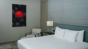 Habitación de hotel con cama, una silla, una mesita de noche con lámpara y una obra de arte en la pared.
