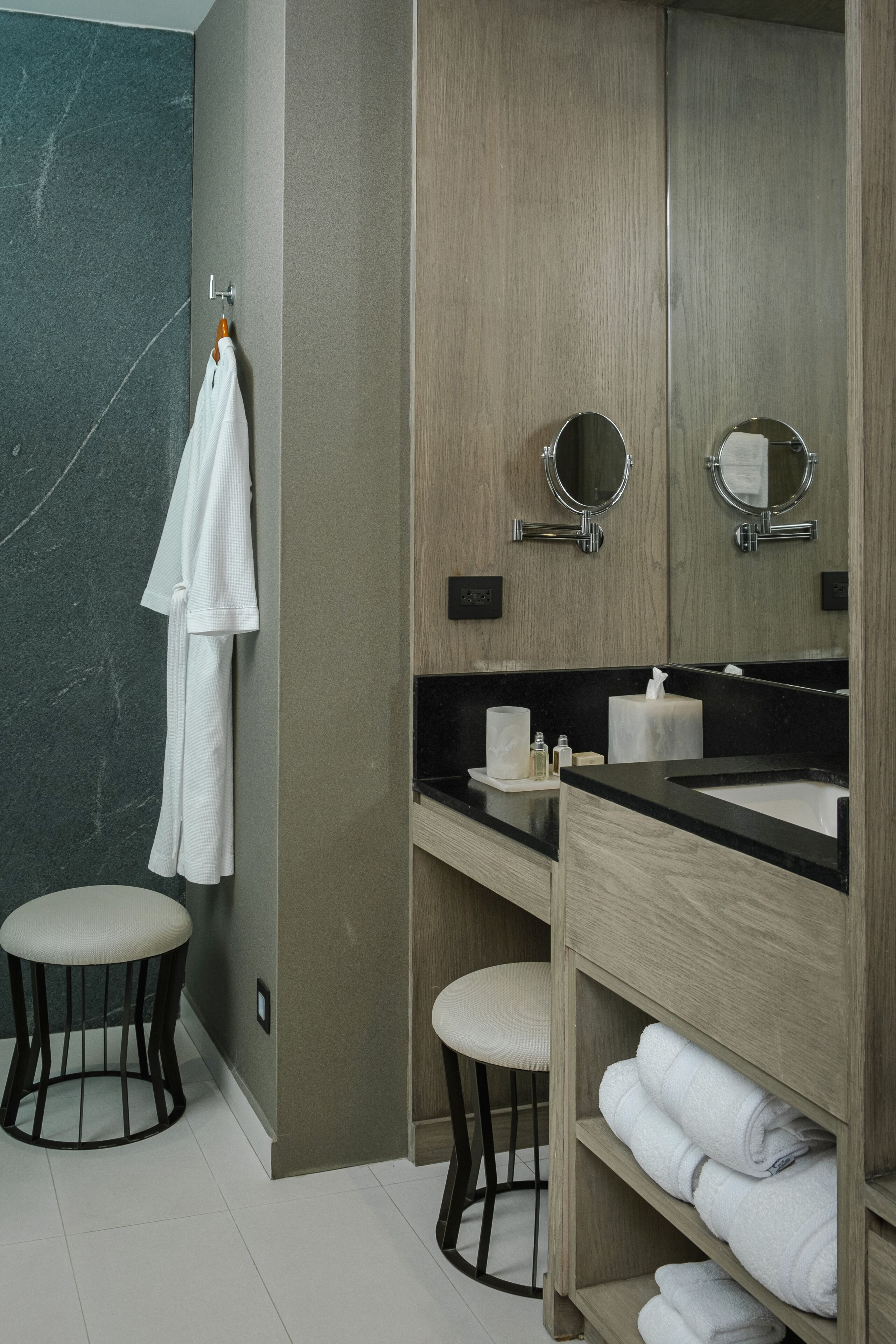 Un baño moderno con pared de mármol, tocador de madera, dos espejos redondos, encimera negra y toallero blanco.