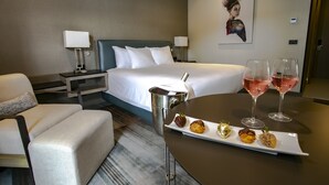 Una habitación de hotel con cama, una mesita de noche con lámpara, una silla, una pequeña mesa con un plato de pasteles y dos copas de vino.