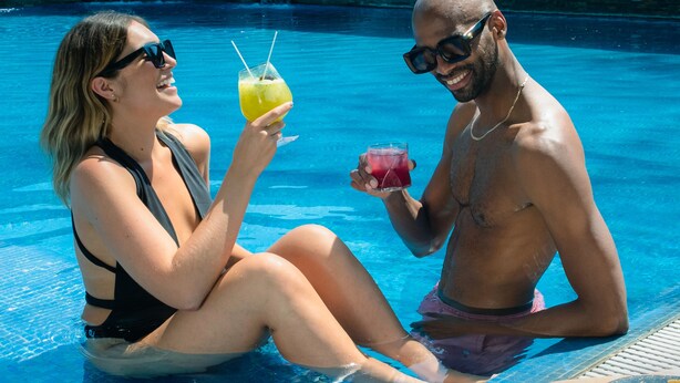 Dos personas sentadas en una piscina con bebidas, piso de baldosas y un muro de piedra al fondo.