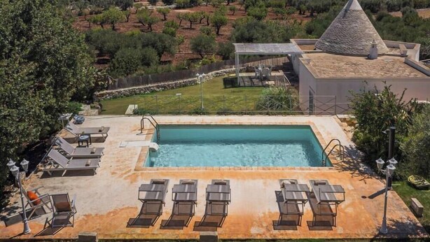 Una piscina rectangular con sillas de descanso y una pérgola, rodeada de un jardín ajardinado y un edificio con techo cónico.
