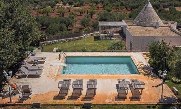A rectangular pool with lounge chairs and a pergola, surrounded by a landscaped garden and a building with a conical roof.