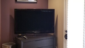 Ein Zimmer mit einem Flachbildfernseher auf einer dunklen Holzkommode, einem kleinen Mülleimer und einer Tür mit weißem Rahmen.