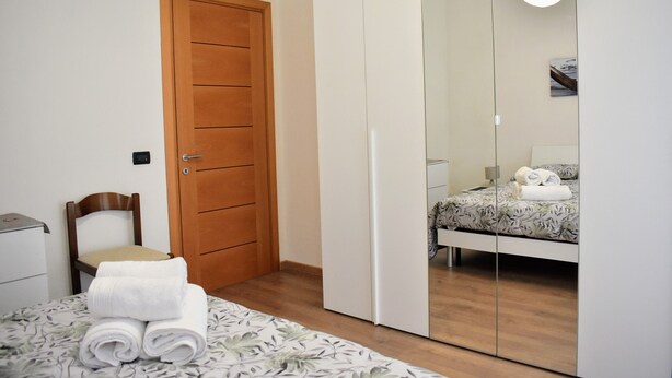 A bedroom with a wooden door, a bed with a floral bedspread, a chair, and a mirror reflecting the bed and a towel. - Your home near Venice - Free Parking (Venezia)