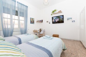 A bedroom with two beds, a desk, a chair, a TV, and a shelf with plants.