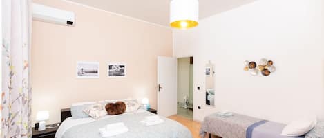 A bedroom with a bed, a bedside table, a nightstand, a bench, a hanging light fixture, and a wall with pictures and decorative items.