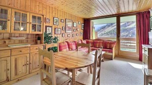 Interior de una cabaña de madera con una mesa de comedor, sillas, un sofá y una cocina con armarios y cuadros en las paredes.