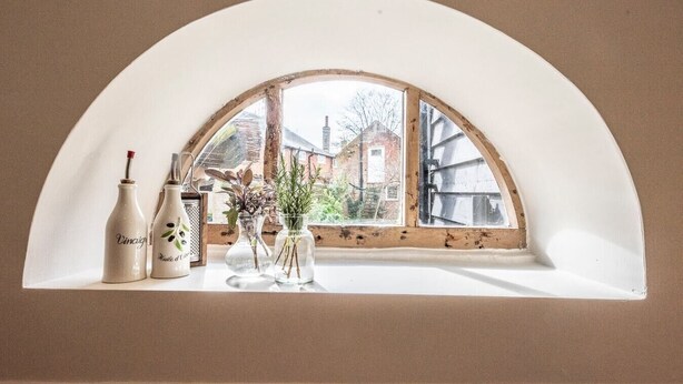 Uma janela com arco branco, pela qual se vê um edifício de tijolos e vegetação.