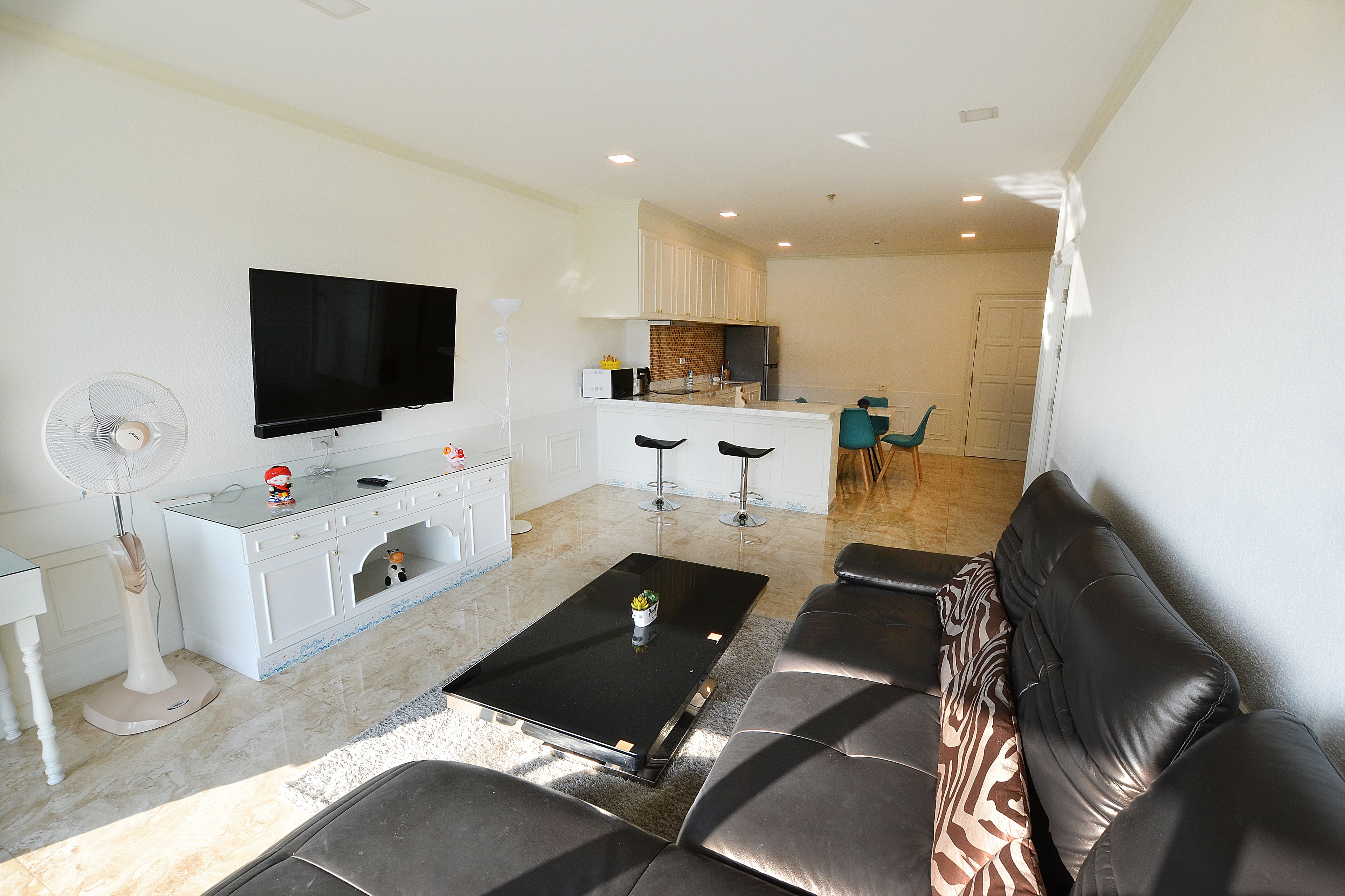 Una sala moderna con una televisión de pantalla plana, una mesa de centro y un sofá. Se observa una zona de cocina con un refrigerador y una mesa de comedor con sillas.
