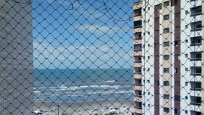 Blick von einem Balkon mit einem Maschendrahtzaun auf Strand und Straße mit Fahrzeugen.