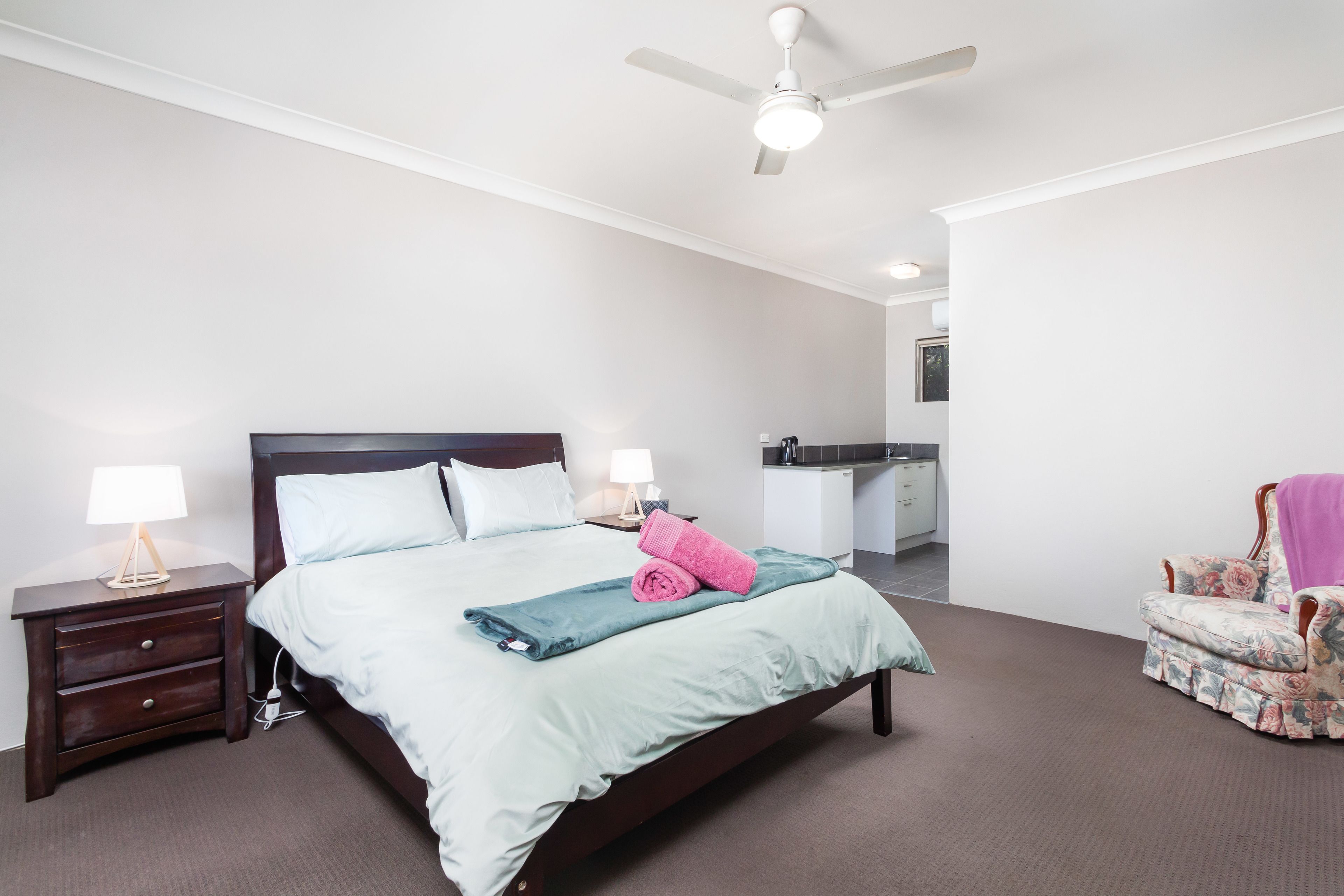 Une chambre à coucher avec un lit, des tables de chevet, une chaise, un ventilateur de plafond et, au fond, une petite cuisine.
