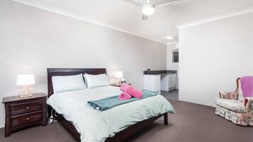 Une chambre à coucher avec un lit, des tables de chevet, une chaise, un ventilateur de plafond et, au fond, une petite cuisine.