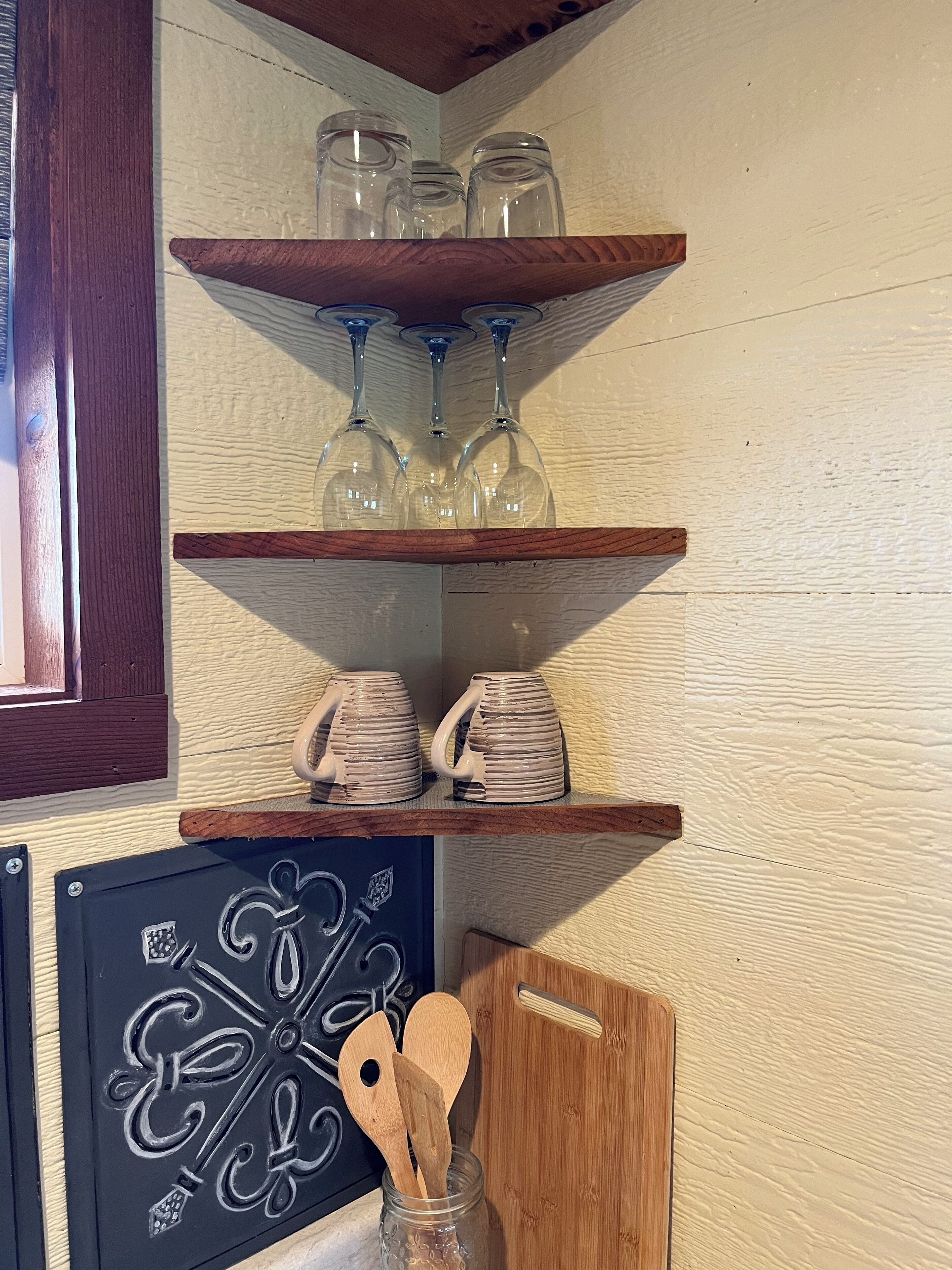 Eine Küchenecke mit Holzböden, die Glaswaren und Keramiktassen beherbergen, einer dekorativen schwarzen Metallplatte und Holzkochern in einem Glas.