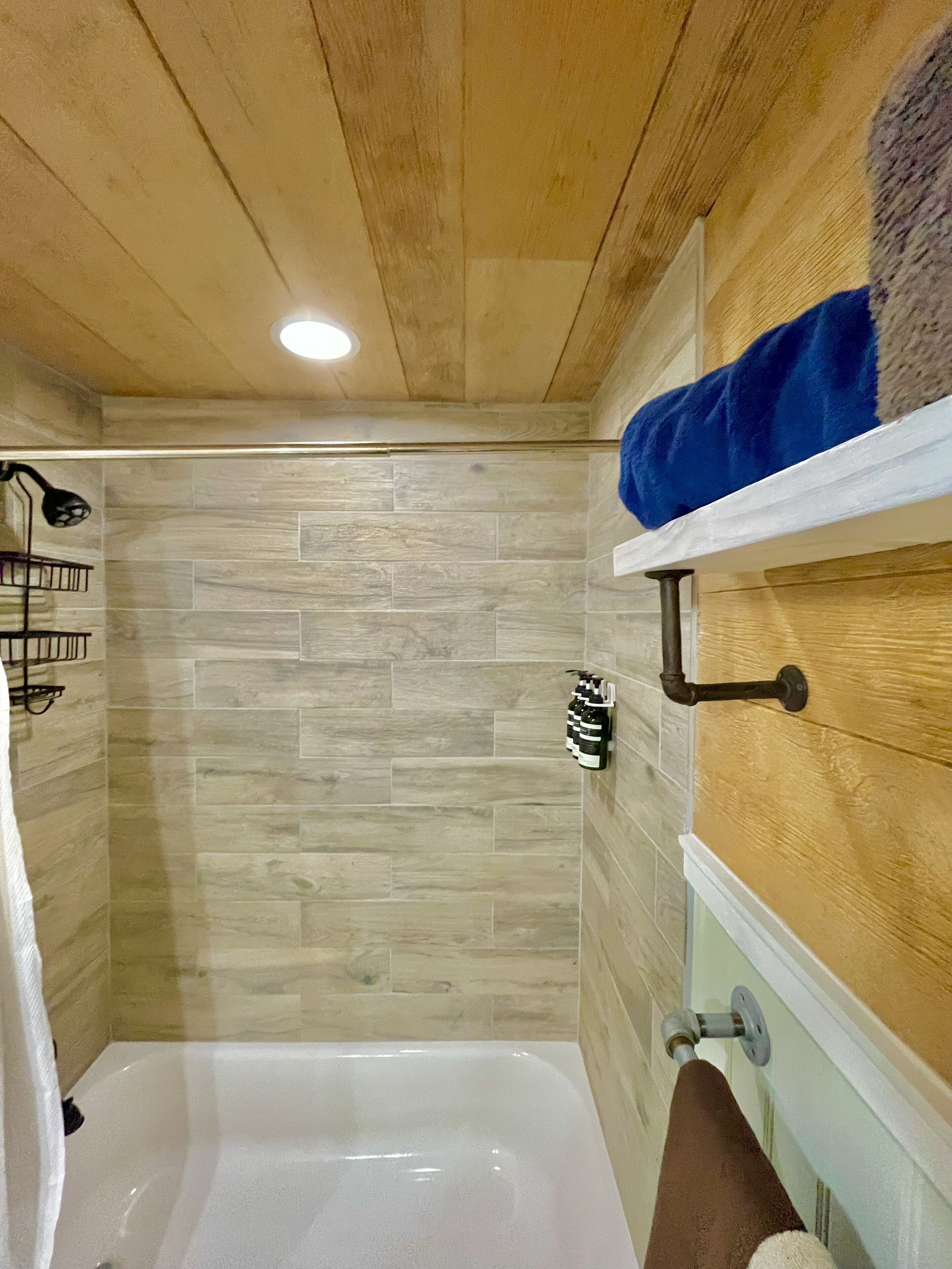 Un baño con techo de madera, zona de ducha con paredes de azulejos, una bañera blanca y un perchero con toallas.