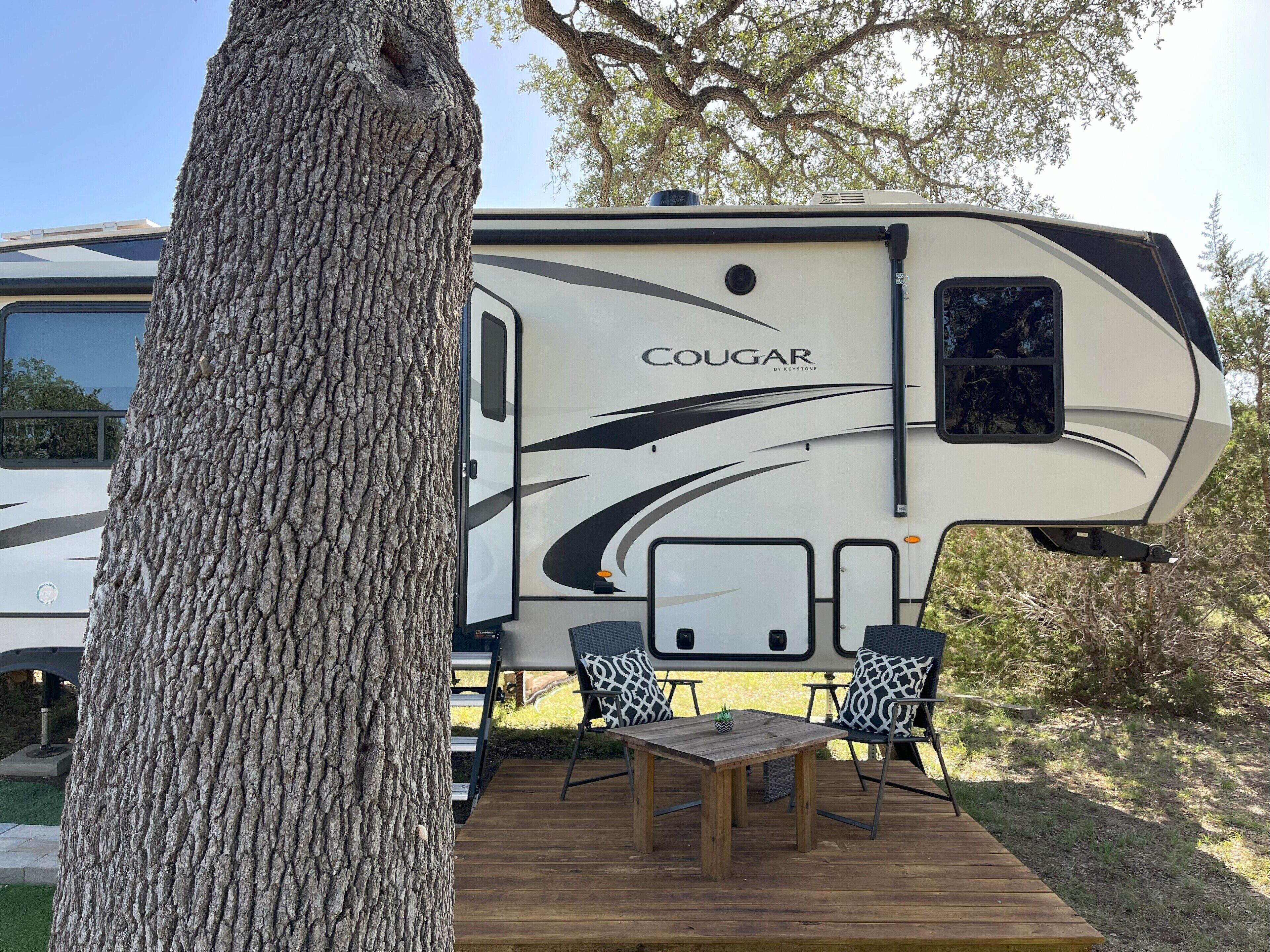 A recreational vehicle with a 'COUGAR' logo, a deck with a table and chairs, and a tree in the foreground.
