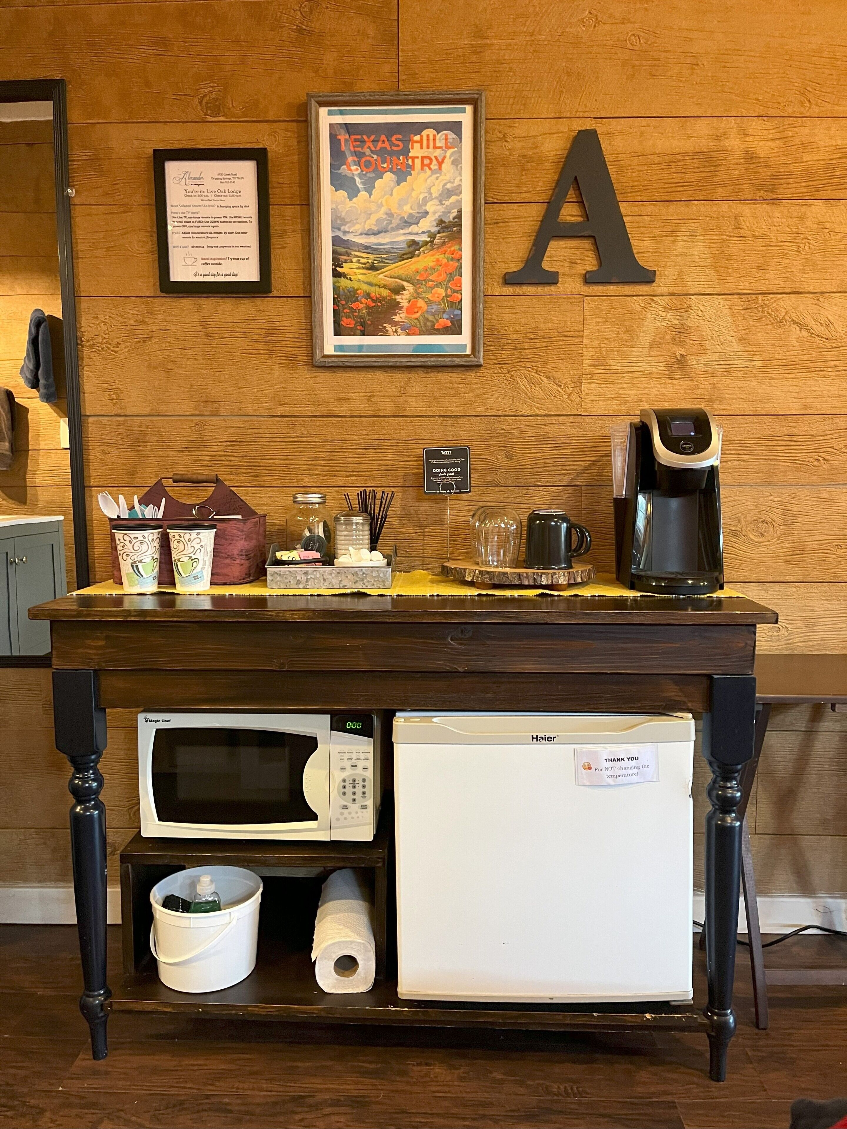A kitchen counter with a coffee maker, microwave, and refrigerator. There is a framed picture of a landscape and a large letter 'A' on the wall.