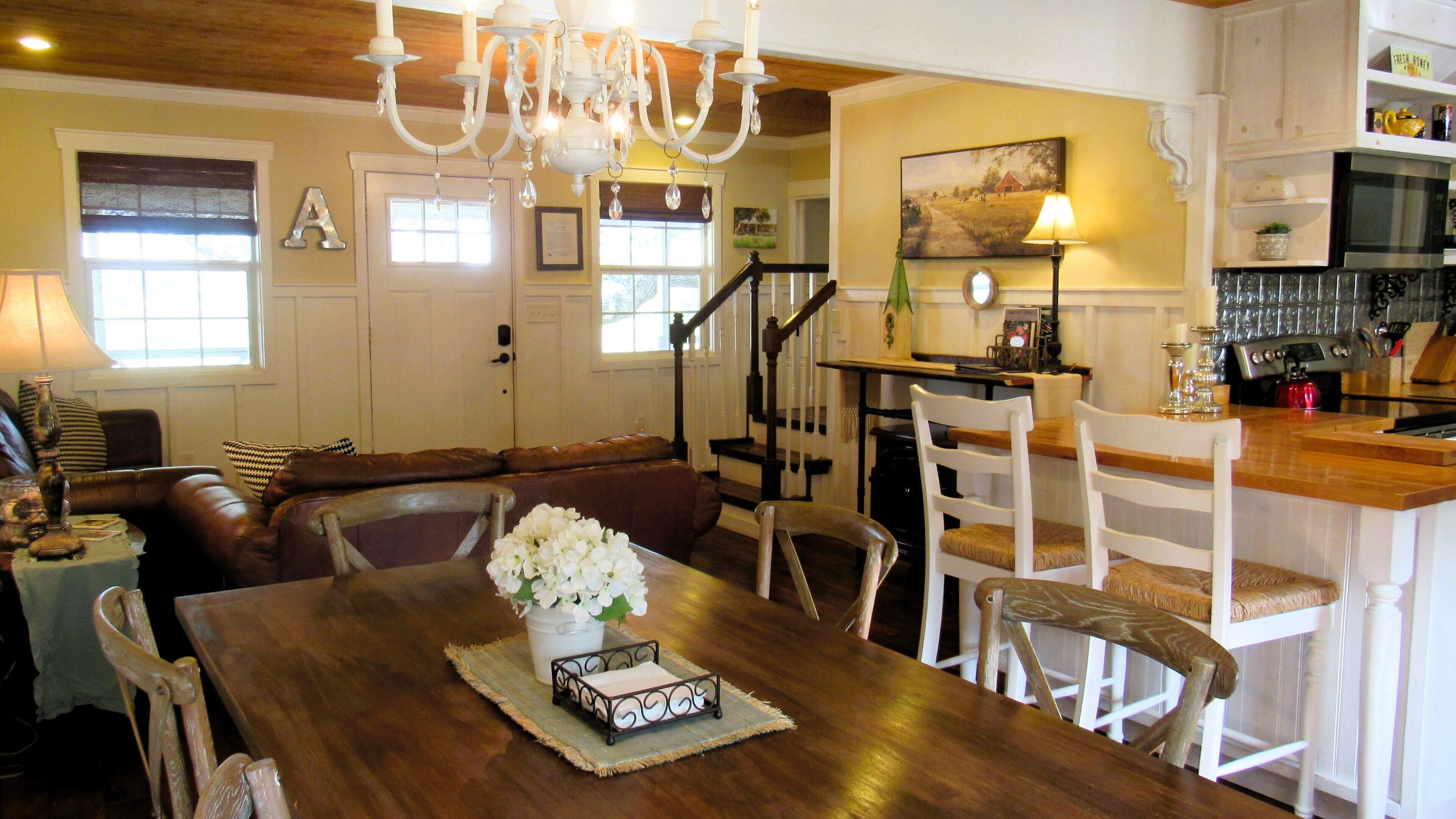 A dining area with a wooden table, white chairs, a chandelier, and a staircase leading to another room.
