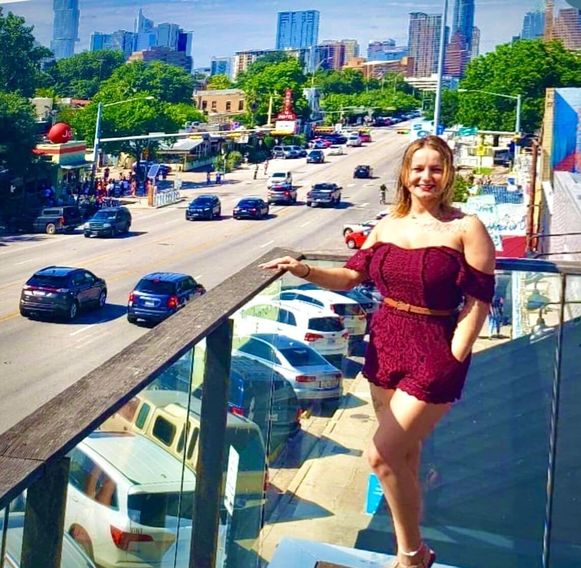 Une femme se tient sur un balcon donnant sur une rue animée de la ville, avec des voitures et des immeubles.