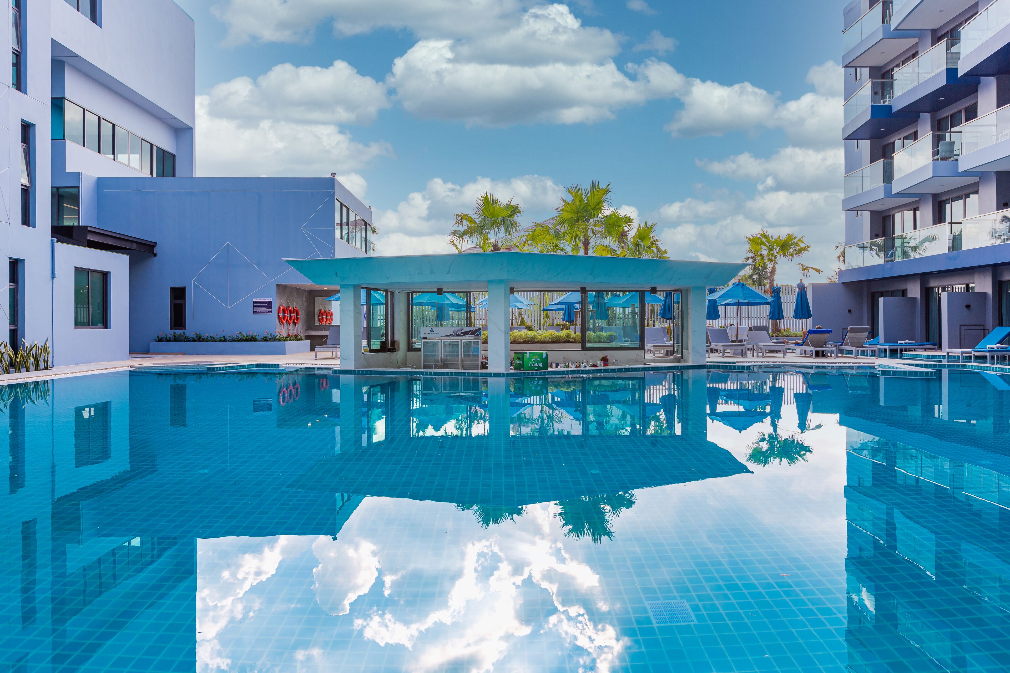 A modern hotel with a large swimming pool, palm trees, and a clear blue sky.