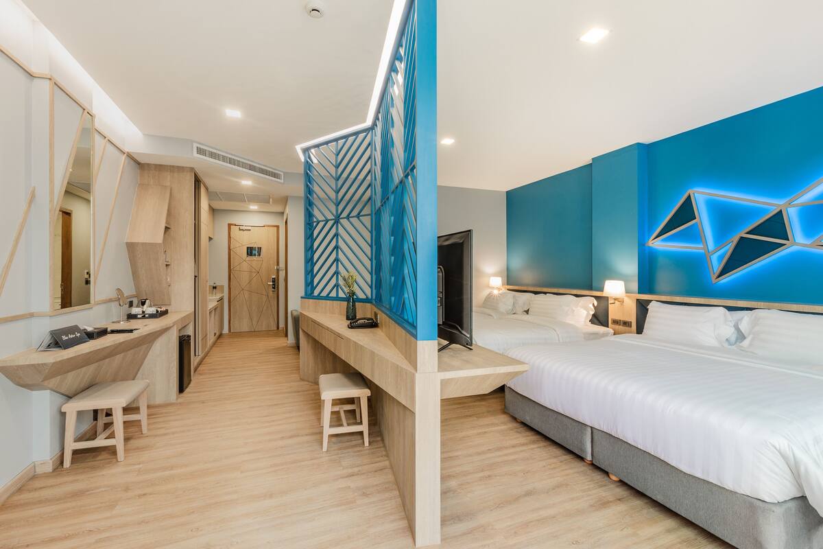A modern hotel room with a wooden desk, two single beds, a large mirror, and a geometric light fixture on the wall.