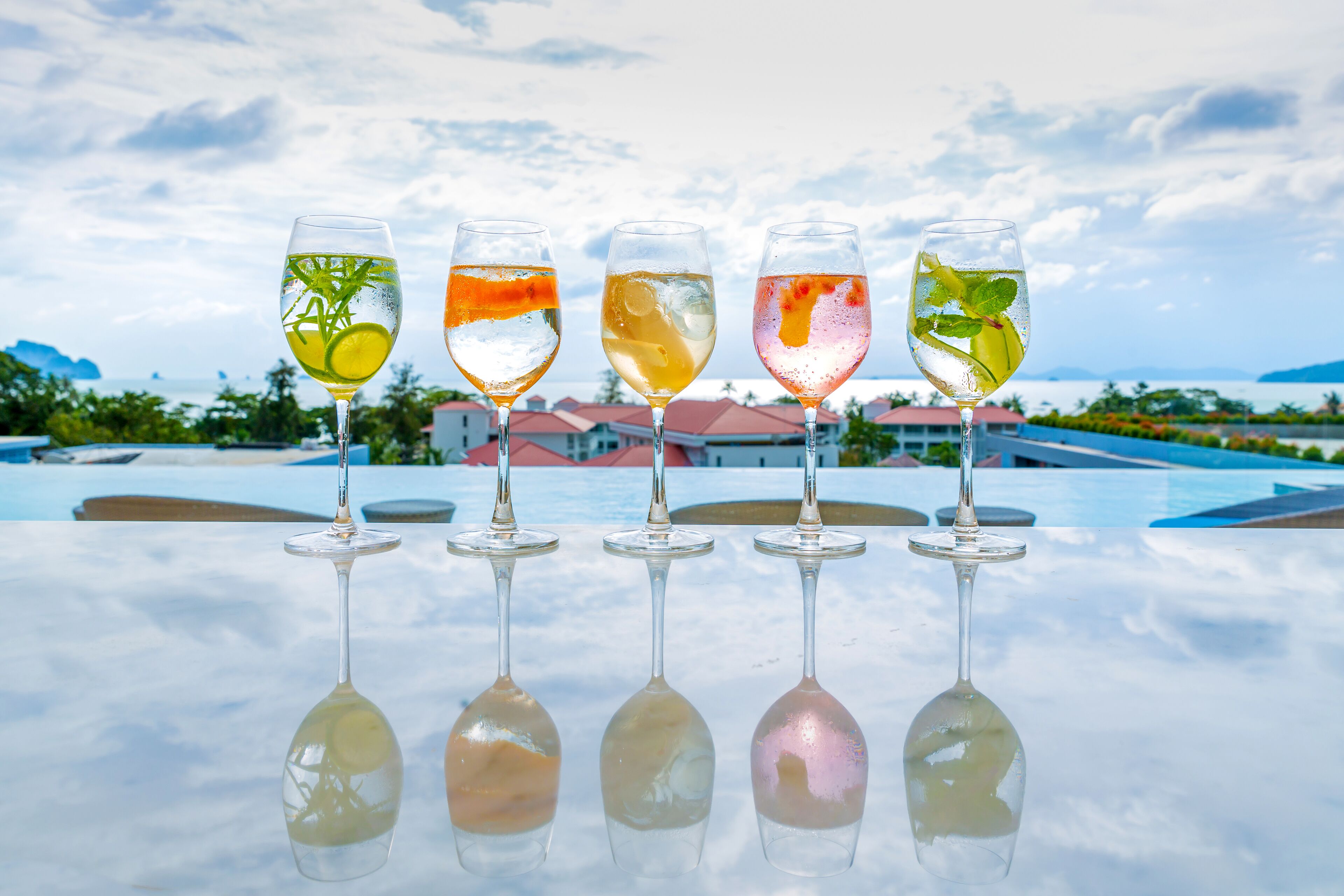 Five glasses of varying colored drinks on a reflective surface with a view of a resort and buildings in the background.