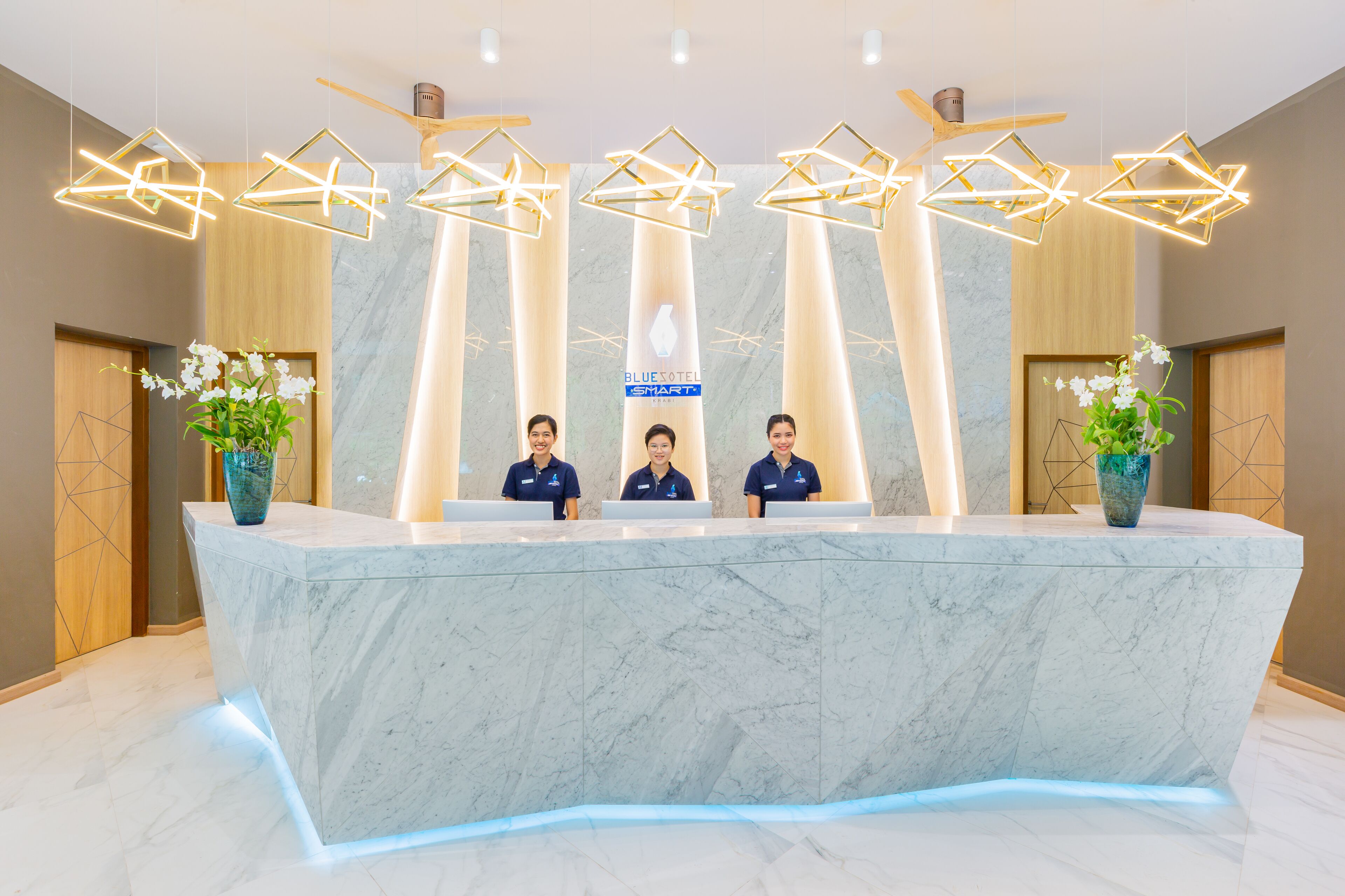 Un mostrador de recepción con tres empleados, una encimera de mármol, luces de techo geométricas y plantas en macetas.
