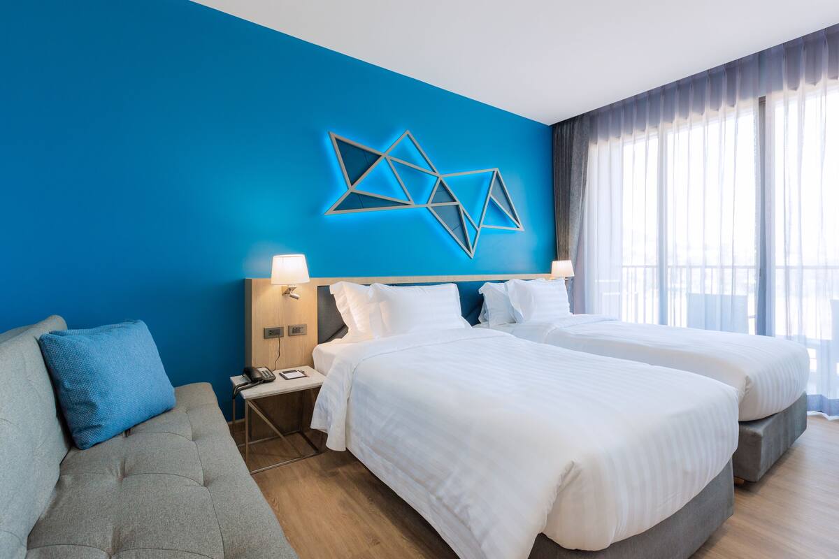 A hotel room with two beds, a sofa, a blue accent wall, and a geometric light fixture.