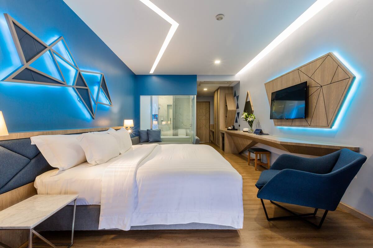 A modern hotel room with a large bed, a desk, a chair, and a TV mounted on the wall.