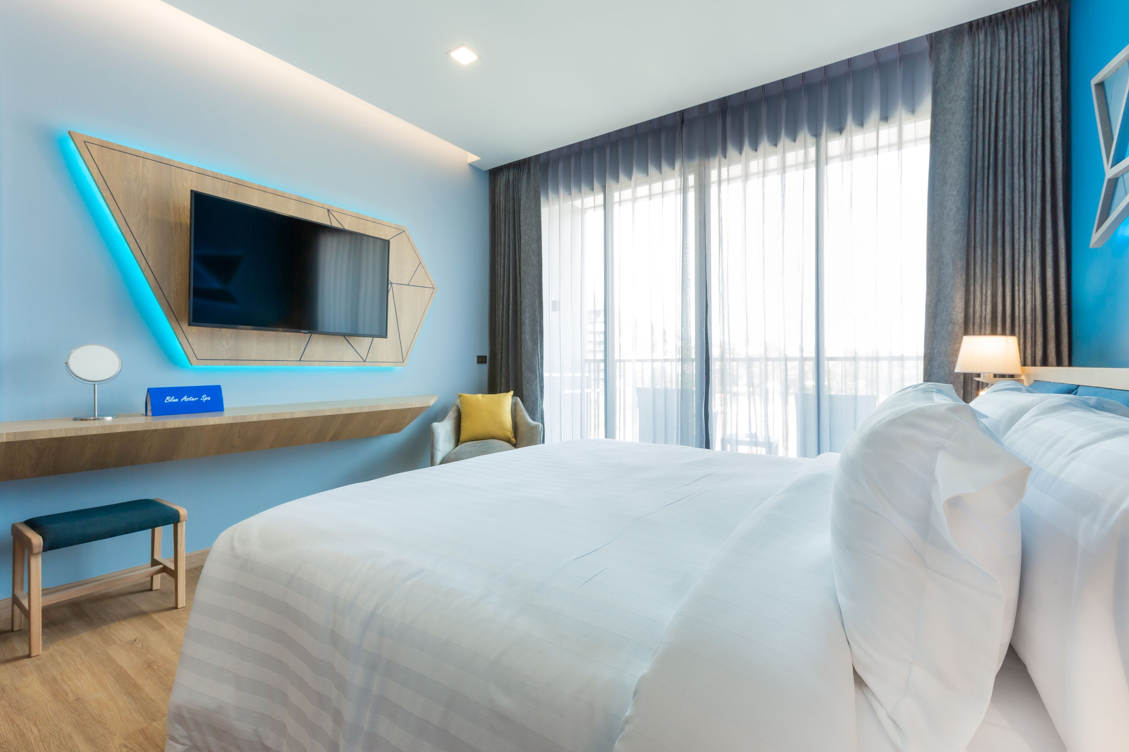 A modern hotel room with a large bed, a TV mounted on the wall, a desk with a chair, and a window with curtains.