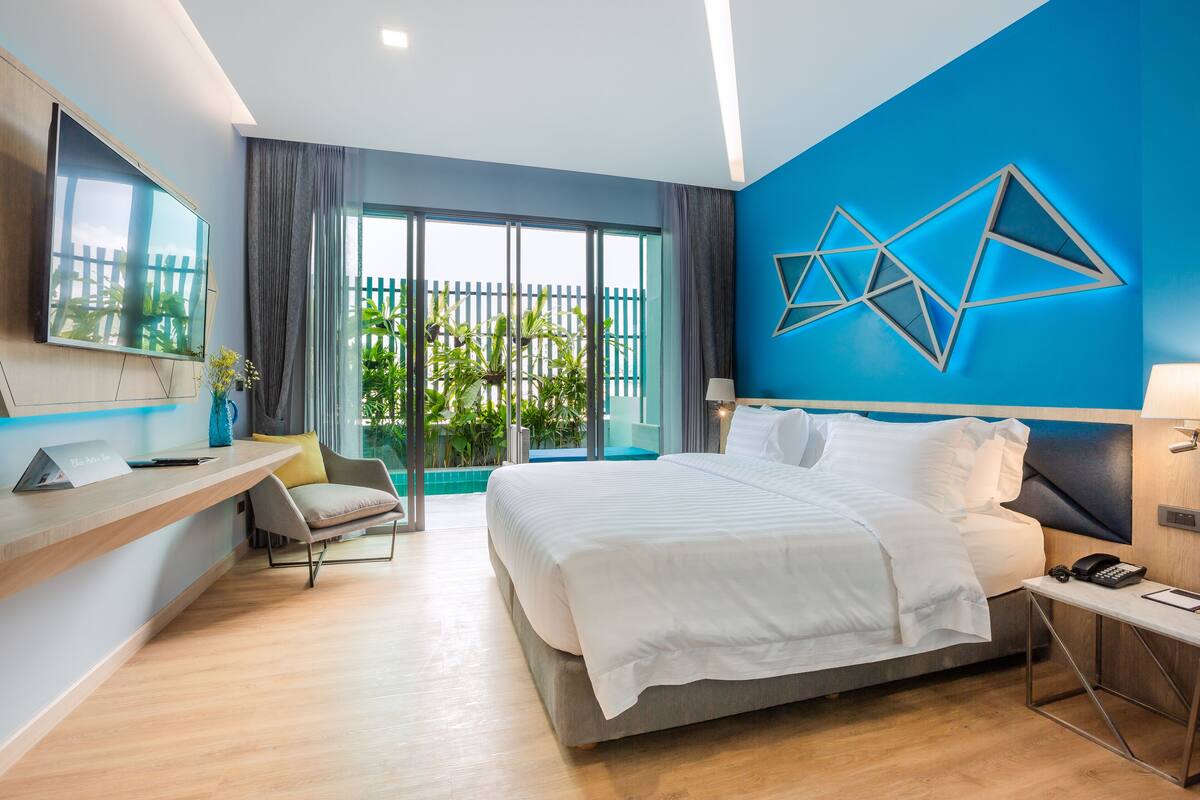 A modern hotel room with a large bed, a desk with a laptop, a chair, and a view of a balcony with plants.