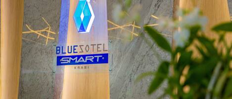 Un letrero para Bluesotel Smart con un logotipo geométrico y el nombre 'KARBI' debajo, montado sobre una estructura de madera con piso de mármol.