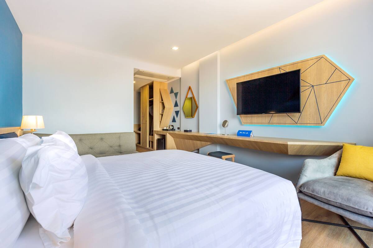A modern hotel room with a large bed, a desk, a TV mounted on the wall, and a seating area with cushions.