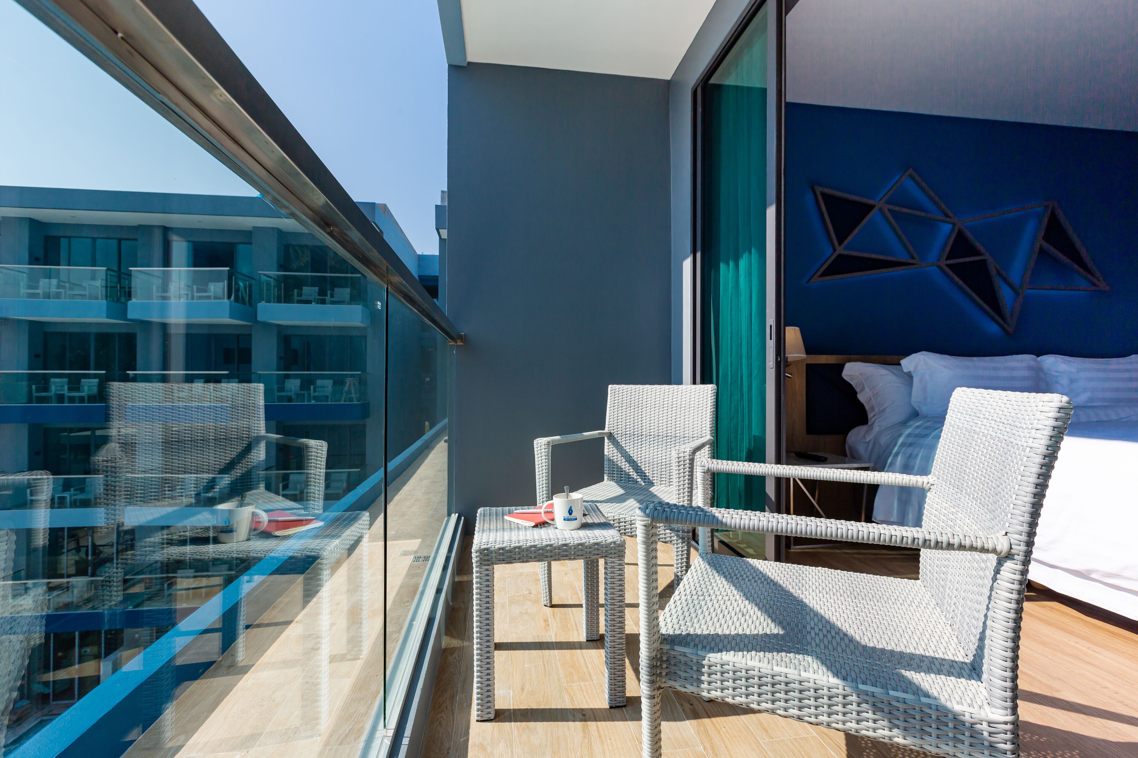Un balcon avec des fauteuils en osier, une petite table et un lit adossé à un mur bleu, décoré de motifs géométriques.