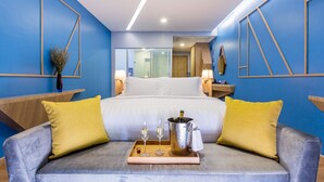 A modern hotel room with a bed, a grey sofa with yellow cushions, a tray with two glasses and a champagne bottle, and a large mirror.