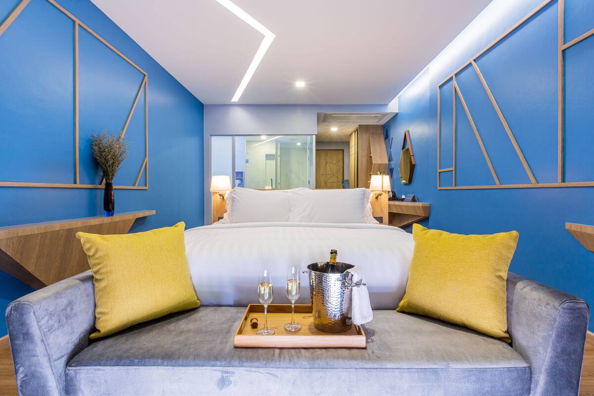 A modern hotel room with a bed, a grey sofa with yellow cushions, a tray with two glasses and a champagne bottle, and a large mirror.