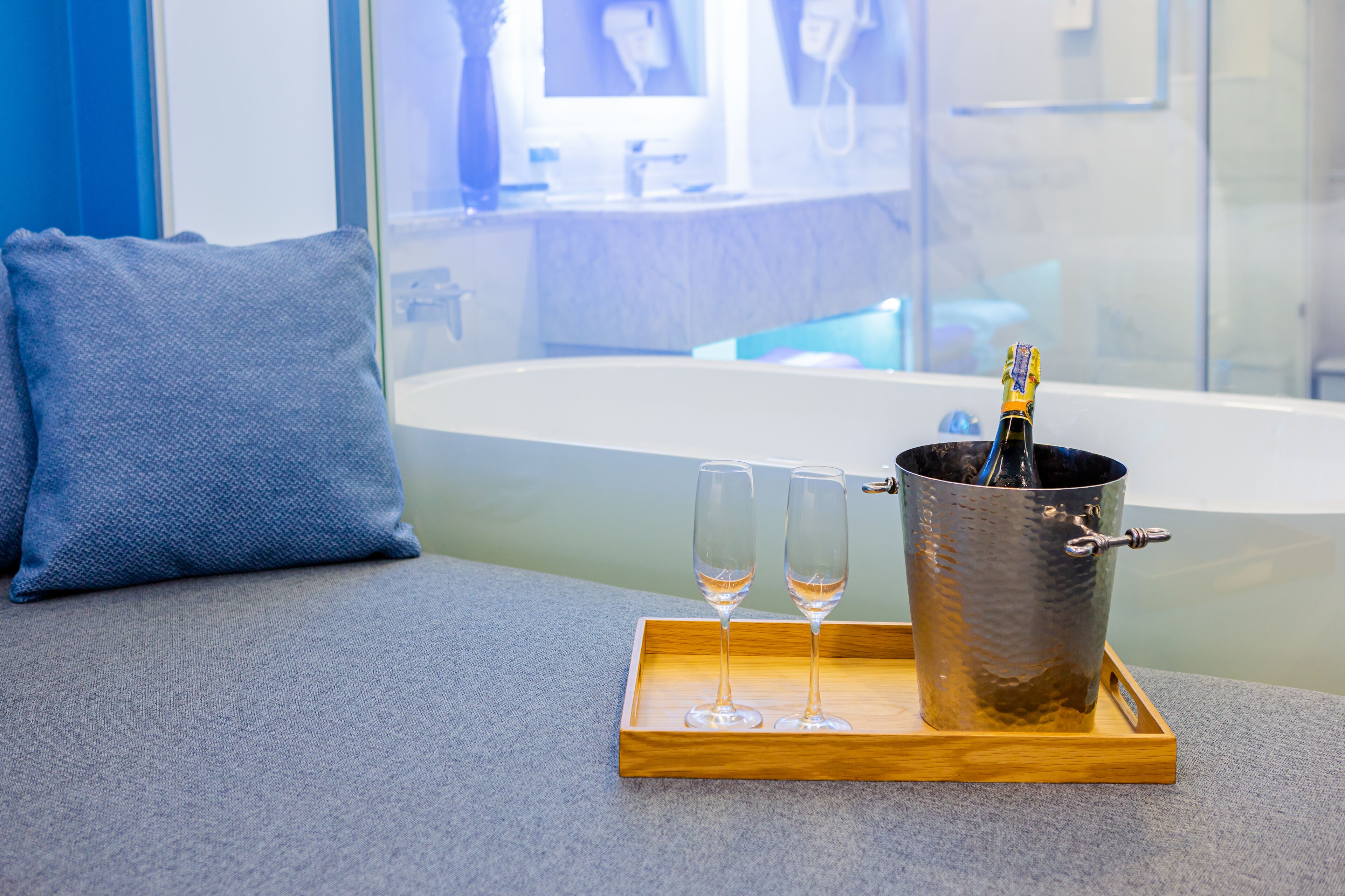 A wooden tray with two glasses and a champagne bottle on a gray sofa in a modern bathroom.