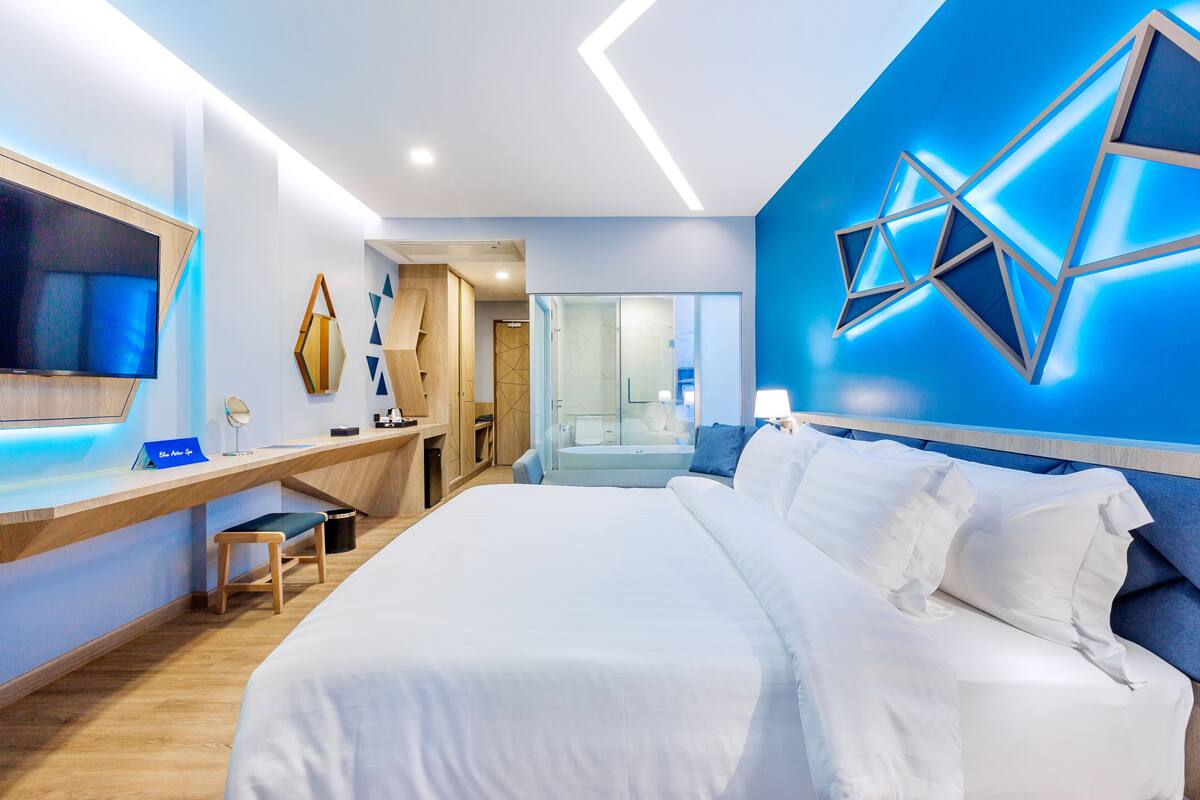 A modern hotel room with a large bed, a desk with a flat-screen TV, and a bathroom visible through an open door.