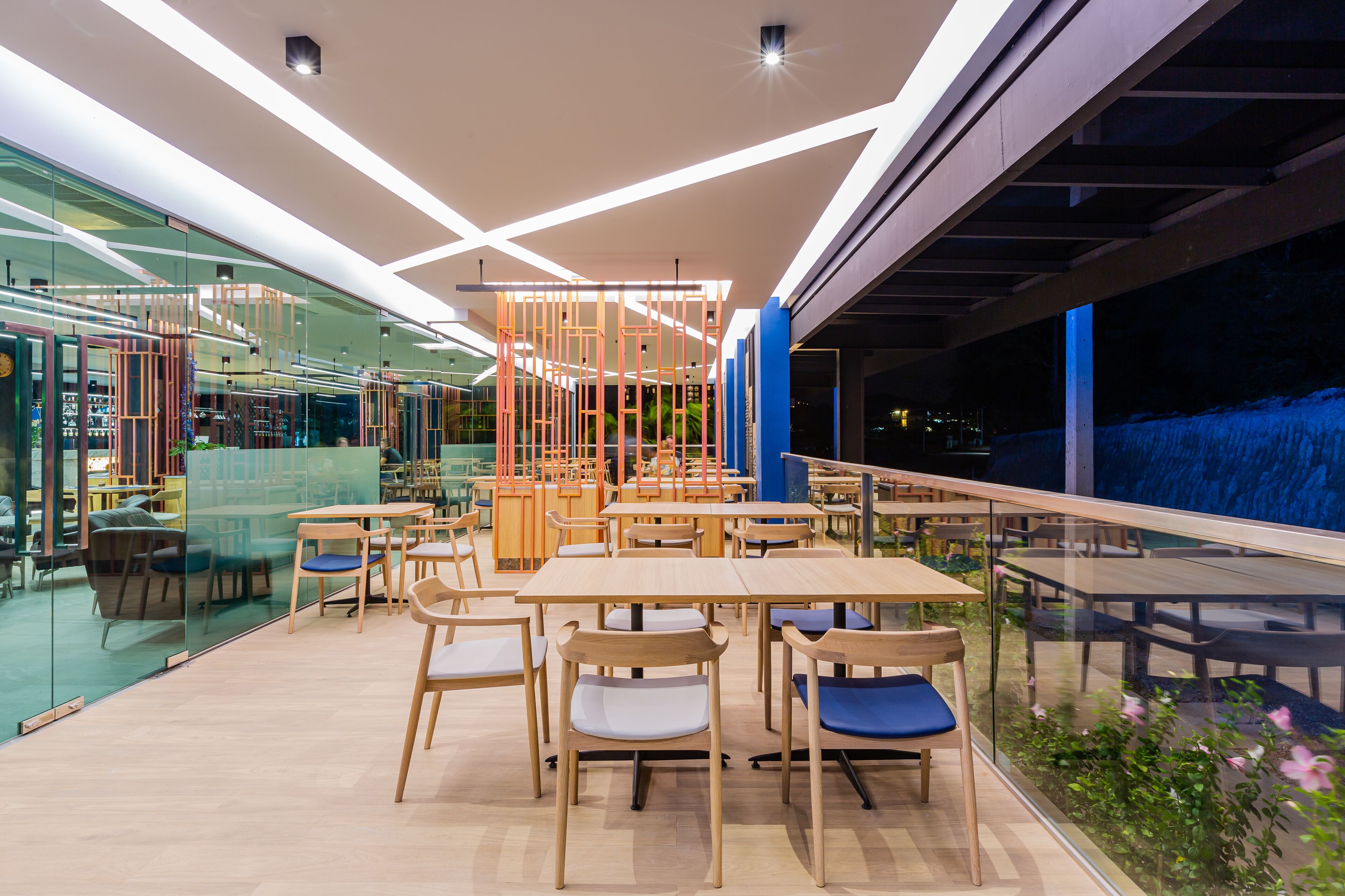 A modern indoor dining area with wooden tables and chairs, glass partitions, and a view of the outside.