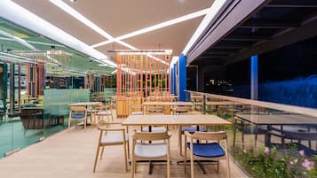 Een moderne, overdekte eetruimte met houten tafels en stoelen, glazen scheidingswanden en uitzicht op buiten.