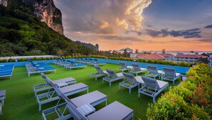 A rooftop pool area with lounge chairs and tables, surrounded by lush greenery and a mountainous backdrop.